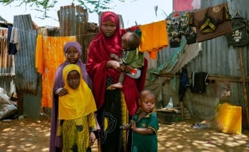 A women stands with four children, her baby in her arms. In front of a corrugated iron wall and clothing line.