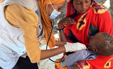 A health worker wearing a face mask and gloves examines a young child with a stethoscope while the child's mother holds them in her arms. The consultation takes place inside a temporary shelter, with the mother watching closely during the health check.