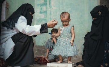 A young girl in a blue dress stands on a scale, with her mother and healthcare worker beside her