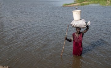 A woman in a red dress wades through waist-deep floodwater, balancing a large sack and bucket on her head while holding a wooden stick for support.