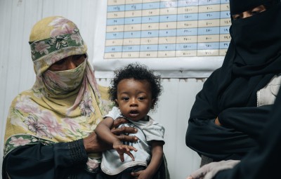 A woman wearing a patterned face covering holds a young child while standing inside a healthcare or community support centre. Another woman wearing a black face veil stands nearby, and a chart is visible on the wall in the background. The child looks toward the camera as the adults participate in what appears to be a health, nutrition, or support service visit. The image highlights maternal and child healthcare support in a community setting.