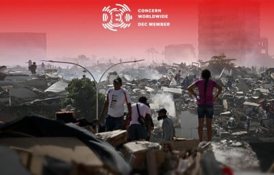 People walk through the aftermath of widespread destruction in a heavily damaged urban area, where collapsed buildings, debris, and smoke stretch across the landscape. A few damaged high-rise buildings remain standing in the background as residents and responders move carefully through the ruins.