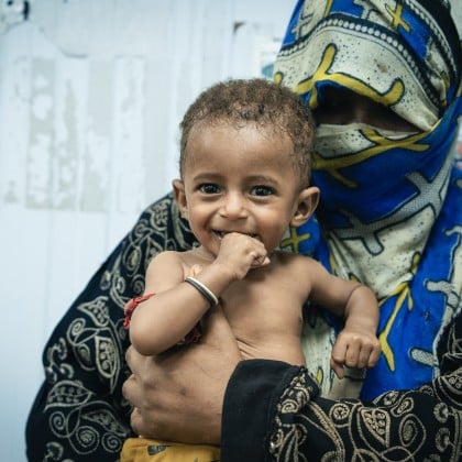 A mother wearing a patterned hijab holds her smiling baby, who is playfully chewing on his hand. The child looks healthy and joyful, a sign of hope amidst adversity.