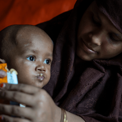 Close-up of a mother wearing a dark headscarf holding and feeding a young child inside a dimly lit shelter or tent. The child is eating from a food sachet held in the mother’s hands.