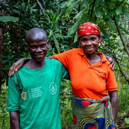 A man in a green T-shirt and a woman in an orange shirt stand arm in arm, smiling towards the camera. They are surrounded by dense green vegetation, with trees and leafy plants filling the background.