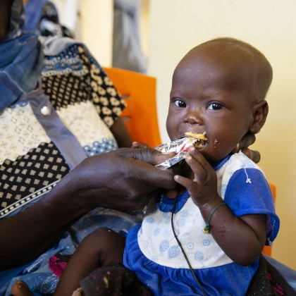 A young child sits on their caregiver’s lap while eating a sachet of ready-to-use therapeutic food (RUTF). The caregiver helps hold the sachet as the child eats during a nutrition programme, with the child looking towards the camera.