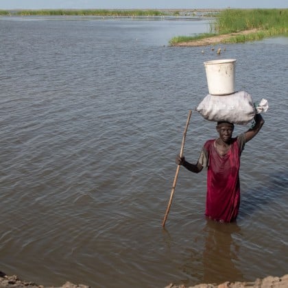 A woman in a red dress wades through waist-deep floodwater, balancing a large sack and bucket on her head while holding a wooden stick for support.