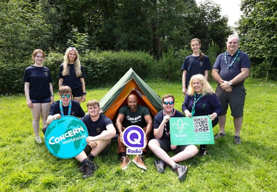 A group of Scouts, Scout leaders, and a Concern Worldwide representative gather around a small tent on a grassy campsite. Several people hold signs featuring the Concern Worldwide logo, Q Radio logo, and Scouts NI logo with a QR code. Surrounded by trees on a sunny day, the group smiles at the camera, highlighting a partnership promoting youth engagement, fundraising, and outdoor adventure.
