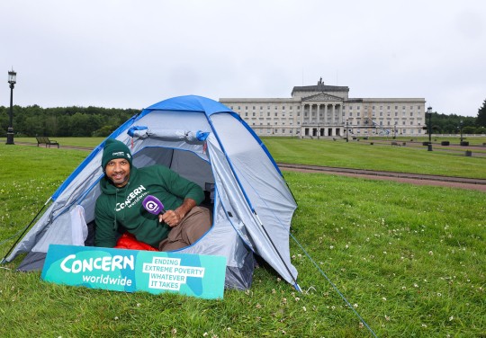 A Concern Worldwide supporter sits inside a blue camping tent on the lawn outside Parliament Buildings at Stormont in Belfast. Wearing a Concern Worldwide hoodie and beanie, they hold a Q Radio microphone beside a sign reading “Concern Worldwide – Ending Extreme Poverty, Whatever It Takes.” 