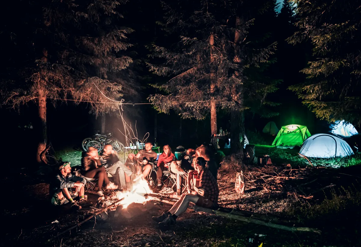 A group of people sit around a glowing campfire at night in a forest clearing, surrounded by tents and tall trees. The scene is warmly lit, with bicycles and camping gear visible in the background.