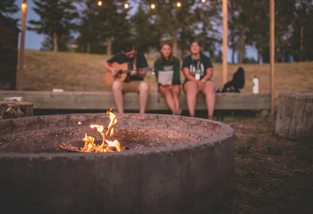A campfire burns in a large circular fire pit in the foreground, while three people sit on a wooden bench in the background. One plays a guitar, and the other two listen and sing along. String lights are hung above, and the setting is surrounded by trees and open space at dusk.