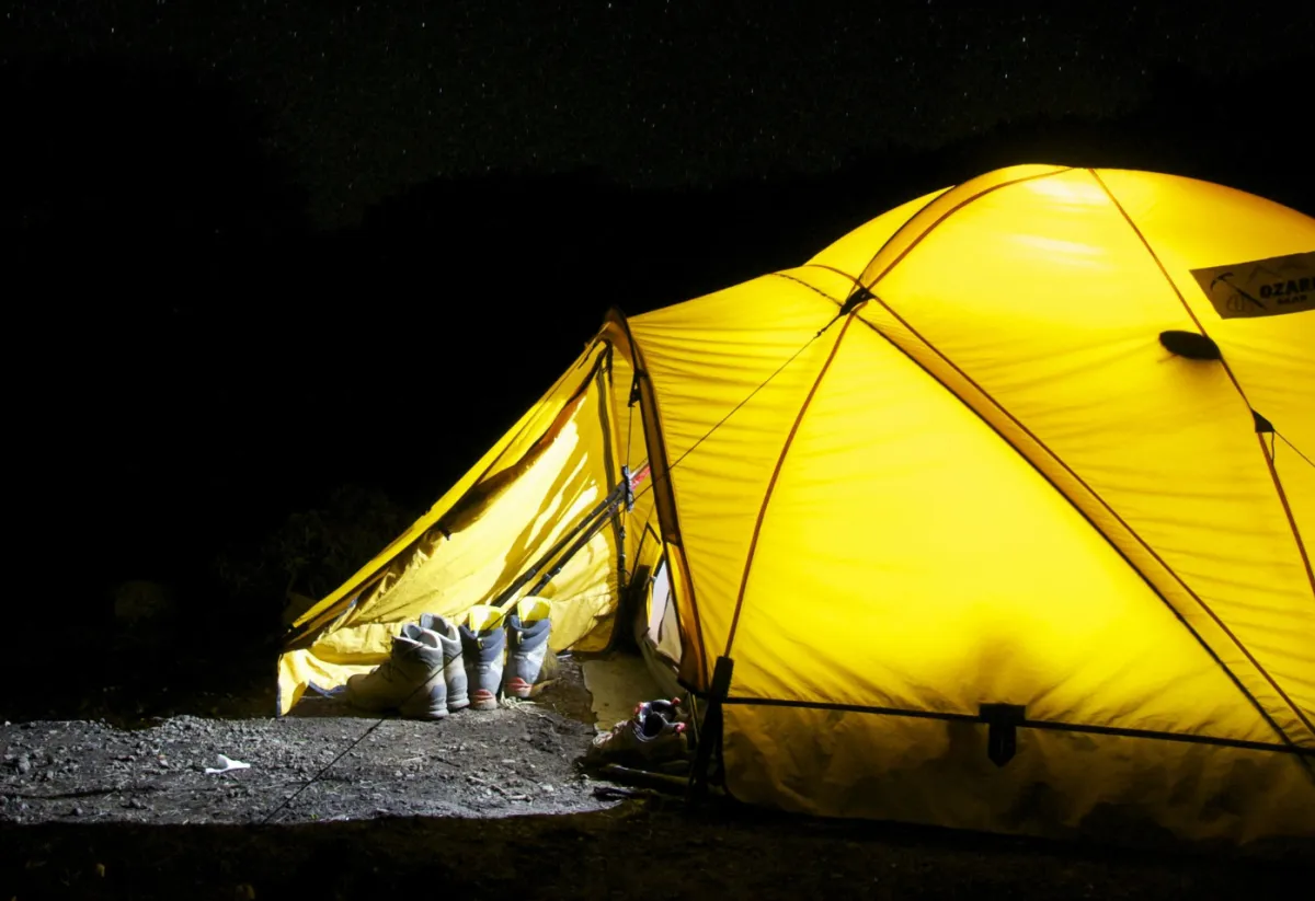 A glowing yellow tent illuminated from within stands in a dark campsite under a star-filled night sky. Hiking boots and trekking poles rest outside the entrance, suggesting a day of adventure.