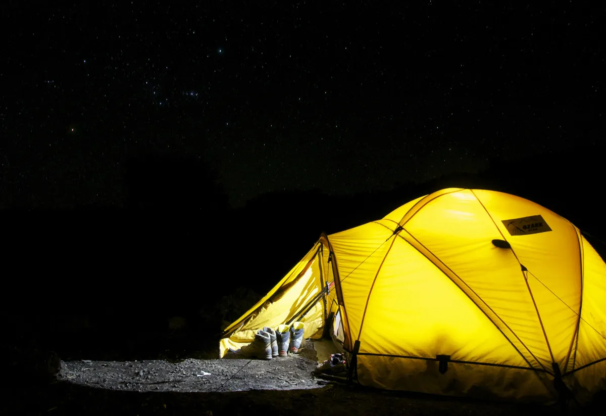 A glowing yellow tent illuminated from within stands in a dark campsite under a star-filled night sky. Hiking boots and trekking poles rest outside the entrance, suggesting a day of adventure.