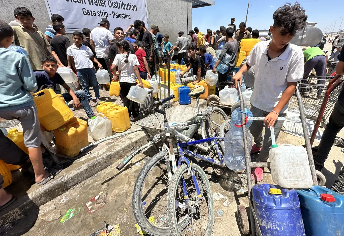 Men and boys fill yellow and white plastic containers with clean water from a truck in a muddy area, while a woman in a patterned black dress stands nearby.