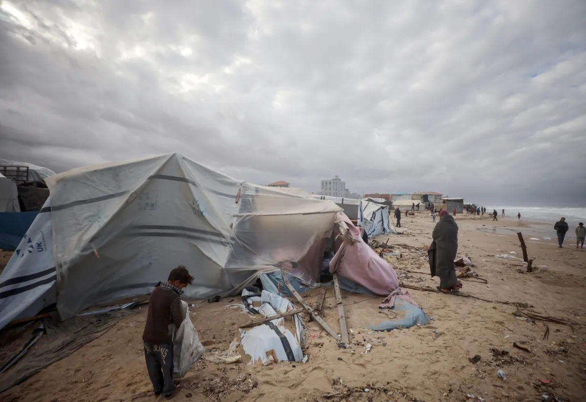 People gather on a beach with debris scattered around. A child and several adults wrapped in blankets walk among the damaged shelters under a cloudy sky.