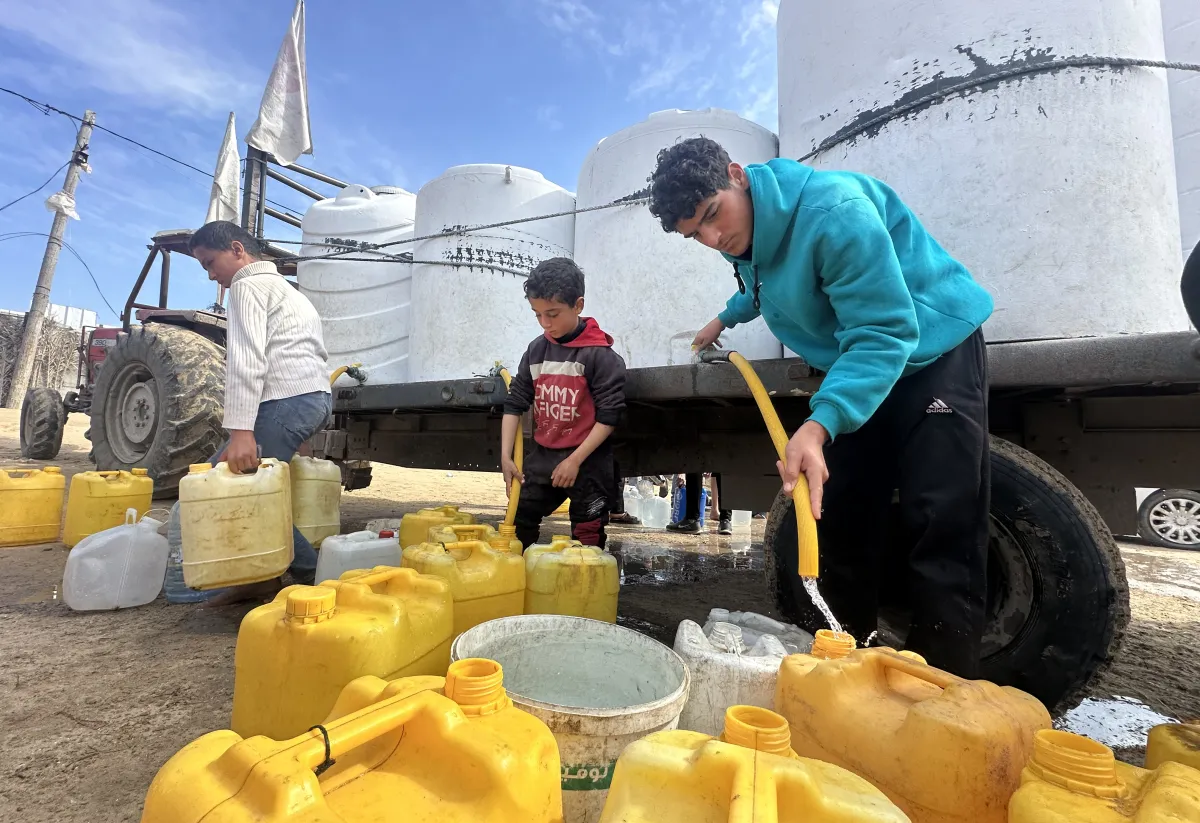 Boys in Gaza collect clean water from large white tanks on a truck, filling yellow 