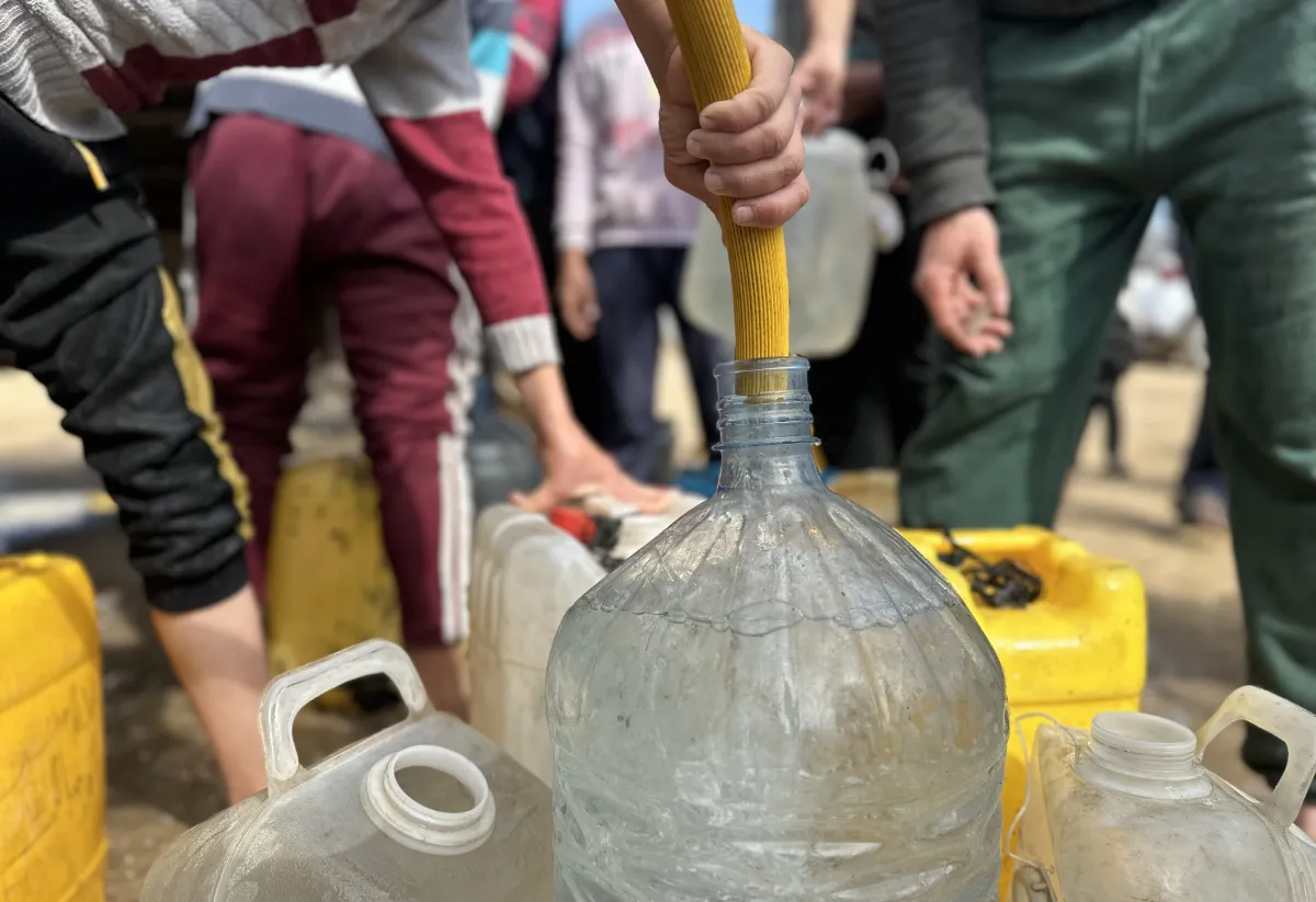 A close-up of a large plastic container being filled with clean water from a yellow hose. Several other jerrycans and people’s hands are visible around, showing the effort of collecting safe water.