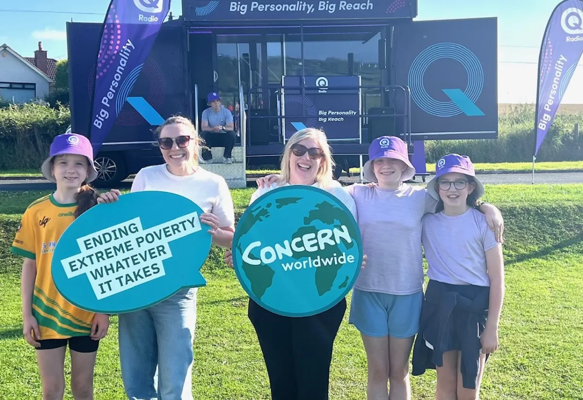 A group of smiling children and two adults stand outdoors holding Concern Worldwide signs, with the Q Radio van in the background.