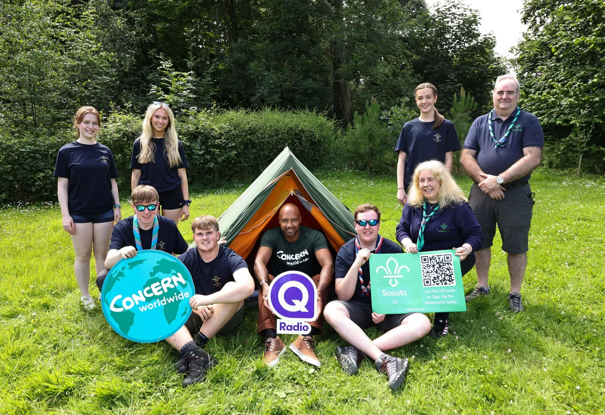 A group of Scouts, Scout leaders, and a Concern Worldwide representative gather around a small tent on a grassy campsite. Several people hold signs featuring the Concern Worldwide logo, Q Radio logo, and Scouts NI logo with a QR code. Surrounded by trees on a sunny day, the group smiles at the camera, highlighting a partnership promoting youth engagement, fundraising, and outdoor adventure.
