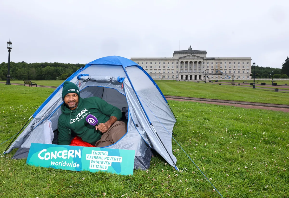 A Concern Worldwide supporter sits inside a blue camping tent on the lawn outside Parliament Buildings at Stormont in Belfast. Wearing a Concern Worldwide hoodie and beanie, they hold a Q Radio microphone beside a sign reading “Concern Worldwide – Ending Extreme Poverty, Whatever It Takes.”