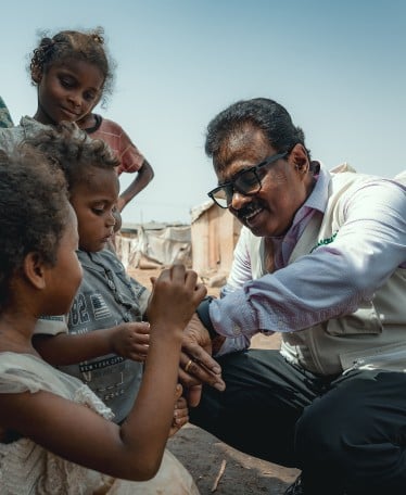 A humanitarian worker from Concern Worldwide kneels down, smiling warmly and engaging with a group of young children in an informal settlement. The children are curiously interacting with his wristwatch. The background shows makeshift shelters under a clear blue sky.