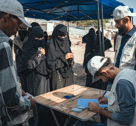 Concern Worldwide staff distribute aid and register community members under a temporary shelter, while women in niqabs and children wait nearby in a humanitarian setting.