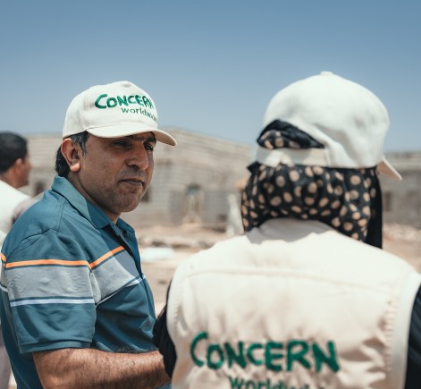 Two Concern Worldwide workers talk amid a humanitarian operation. One wears a branded vest and cap, and the other wears a blue shirt with a Concern cap, with tents and unfinished buildings in the background under a clear sky.