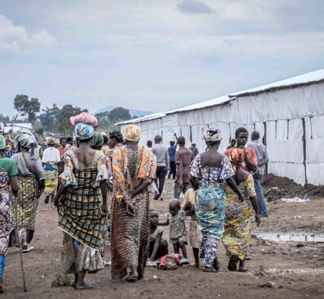 A group of displaced people, mostly women and children in colorful clothing, walk through a settlement lined with temporary shelters constructed from tarpaulin and wood under a cloudy sky.