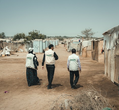 Three Concern Worldwide staff members walk through an informal settlement with makeshift shelters under the hot sun, assessing needs and connecting with the community.