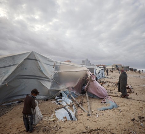 People gather on a beach with debris scattered around. A child and several adults wrapped in blankets walk among the damaged shelters under a cloudy sky.
