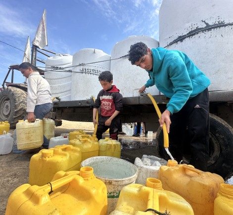Boys in Gaza collect clean water from large white tanks on a truck, filling yellow 