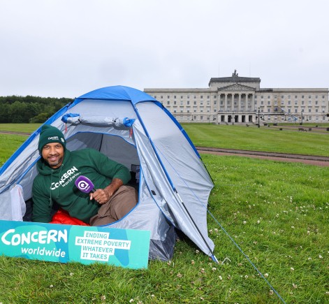 A Concern Worldwide supporter sits inside a blue camping tent on the lawn outside Parliament Buildings at Stormont in Belfast. Wearing a Concern Worldwide hoodie and beanie, they hold a Q Radio microphone beside a sign reading “Concern Worldwide – Ending Extreme Poverty, Whatever It Takes.”