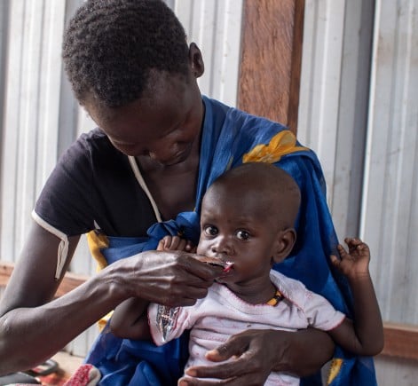 A woman cradles a young child while feeding them a sachet of nutrient-rich food. The child looks directly at the camera as the mother gently supports them in her arms.