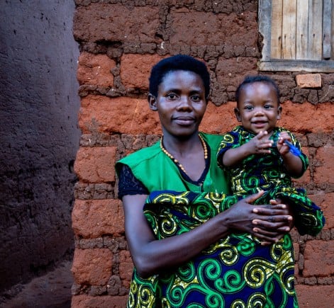 A woman holds a smiling young child outside a home built from mud bricks. Both wear matching green patterned clothing and look toward the camera, while the child smiles and holds a small blue object. A wooden shuttered window is visible behind them.