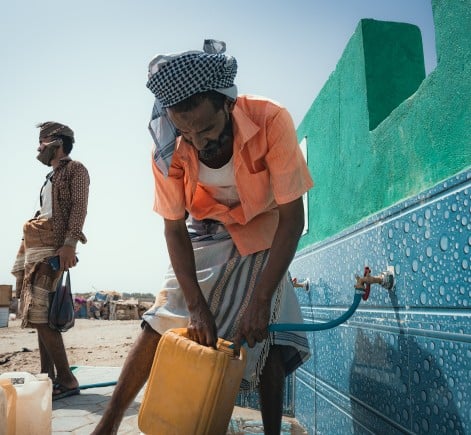 A man fills a yellow jerrycan with water from a public tap connected to a tiled water point. Another man stands nearby holding a bag, while several empty water containers and a motorbike are visible in the background.