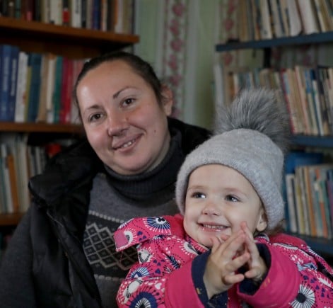 A woman and a young child sit together in a library filled with bookshelves. The child, wearing a pink coat and a grey hat with a pom-pom, smiles with clasped hands while the woman smiles towards the camera.