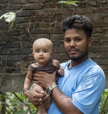 A man in a blue shirt holds a baby in his arms while standing outdoors in front of a brick wall. The baby looks directly at the camera, and both appear calm and composed.