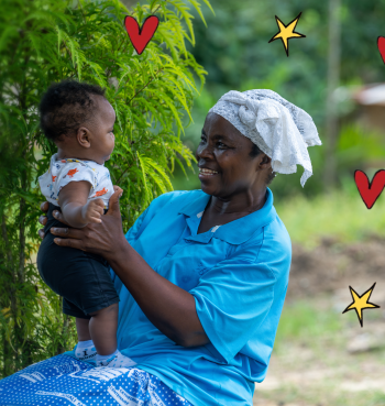 An older woman wearing a blue top and white headscarf sits outdoors holding a baby, smiling as they look at each other. Green foliage surrounds them, with a rural setting and a small building softly blurred in the background.
