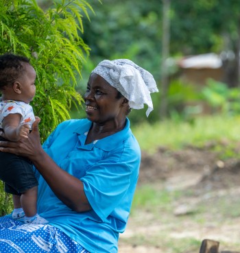 An older woman wearing a blue top and white headscarf sits outdoors holding a baby, smiling as they look at each other. Green foliage surrounds them, with a rural setting and a small building softly blurred in the background.