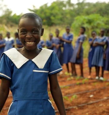 A smiling schoolgirl wearing a blue school uniform stands in the foreground of a school garden. Behind her, a group of students in matching blue uniforms stand together, slightly out of focus. Rows of young plants grow in the red soil around them, with green vegetation in the background. The image conveys education, community, and young people participating in school-based gardening activities.