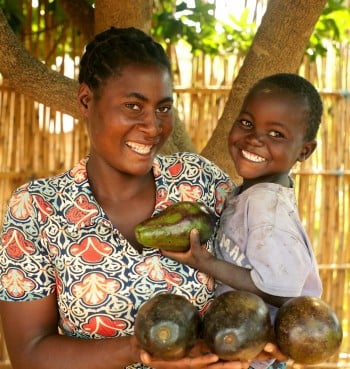 A mother and son smile at the camera holding large, shiny avocados they have grown.