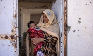 Nasima* (holds her son Rahmin* (2 months old) standing in the doorframe of what was once Sangin&#039;s clinic, bombed out and destroyed. There is currently no healthcare available. Photo: Stefanie Glinski