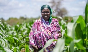 A smiling woman wearing purple stands in a field surrounded by plants.