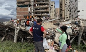 Aid worker and resident stand beside rubble as rescue teams search debris in front of a heavily damaged apartment building.