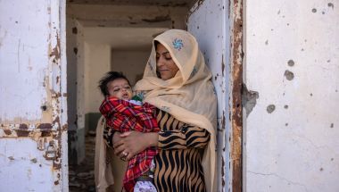 Nasima* (holds her son Rahmin* (2 months old) standing in the doorframe of what was once Sangin&#039;s clinic, bombed out and destroyed. There is currently no healthcare available. Photo: Stefanie Glinski