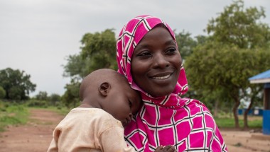 A person wearing a pink and white patterned headscarf holds a child outdoors. In the background, there are trees and a building with a blue roof.