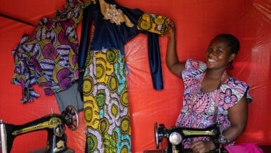 Woman smiling while seated at a sewing machine, holding up a colorful dress with yellow, blue, and green patterns and lace detailing. A second garment with purple and pink designs hangs nearby against a red fabric backdrop