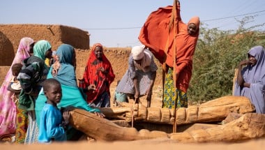Women collect water from a wooden village well. Her orange hijab billows in the wind as she pulls water up from the well. Behind are orange building and a clear, sunny sky.