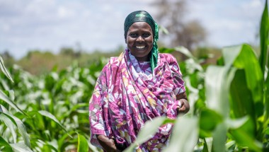 A smiling woman wearing purple stands in a field surrounded by plants.