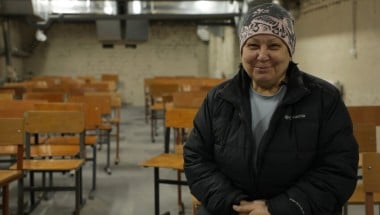 A person wearing a winter jacket and patterned hat sits in a room filled with rows of wooden school chairs. The setting appears to be a basement shelter with exposed brick walls and overhead ventilation ducts.